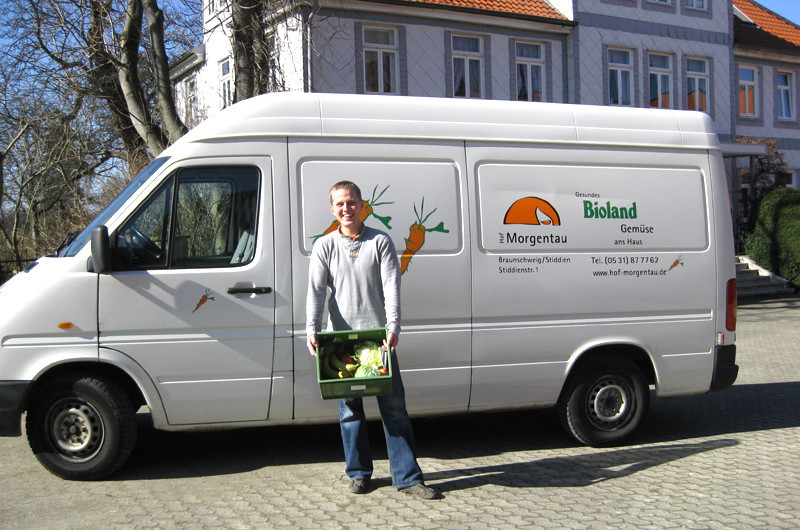 Mitarbeiter mit frisch gepackter Biokiste vor dem Hof-Morgentau-Lieferwagen